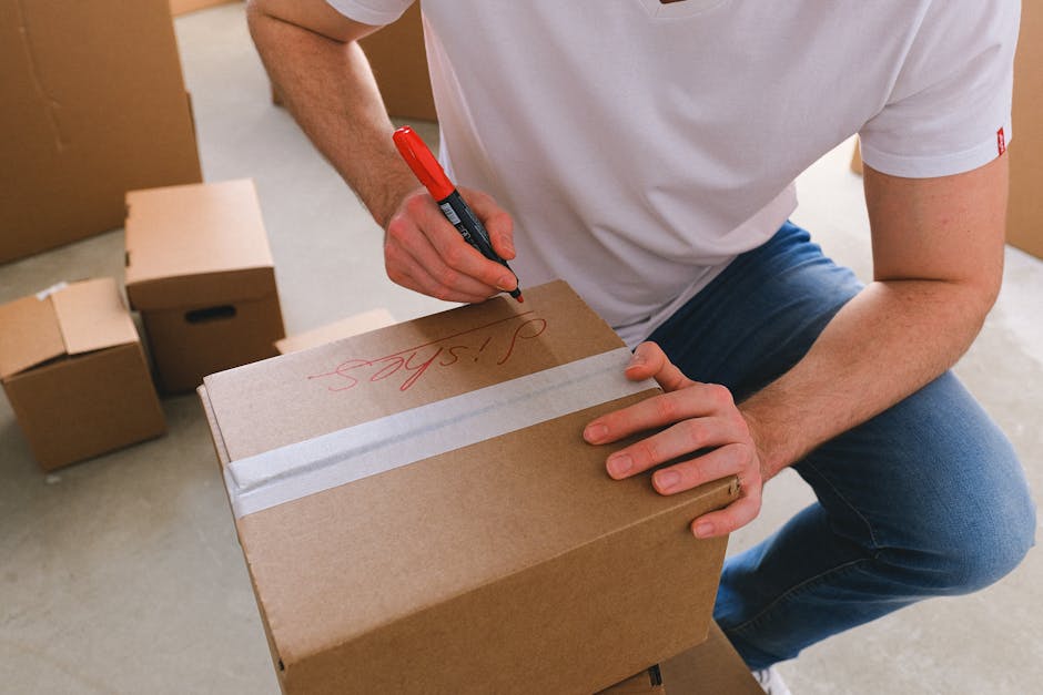 A person wearing a white T-shirt and blue jeans is kneeling indoors, using a red marker to write the word 'Jobs' on a sealed cardboard box with white packing tape. Several other cardboard boxes of var