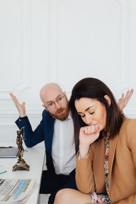 A man with glasses, a beard, and a blue suit shirt appears frustrated, gesturing with both hands raised and palms facing upwards, while seated at a desk inside a room with white panelled walls. Next t