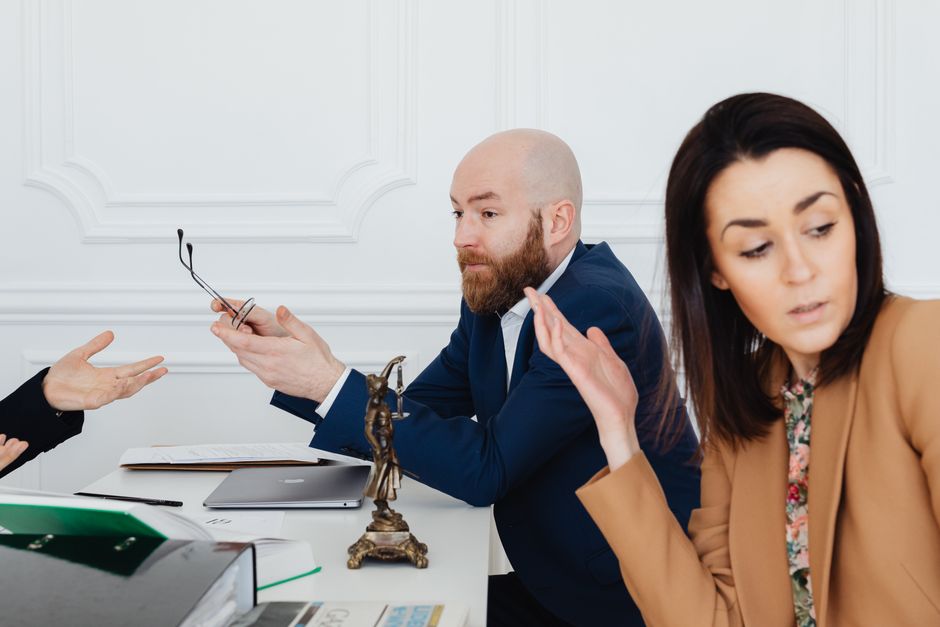 A man with glasses, a beard, and a blue suit shirt appears frustrated, gesturing with both hands raised and palms facing upwards, while seated at a desk inside a room with white panelled walls. Next t