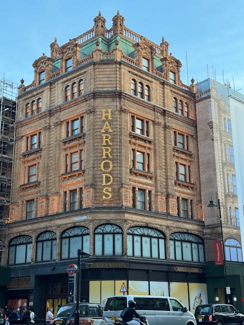 Photograph of a historic multi-story building situated on a busy street near Brompton Road in Knightsbridge, with the name 'HARRODS' displayed vertically on the facade in large gold lettering. The bui