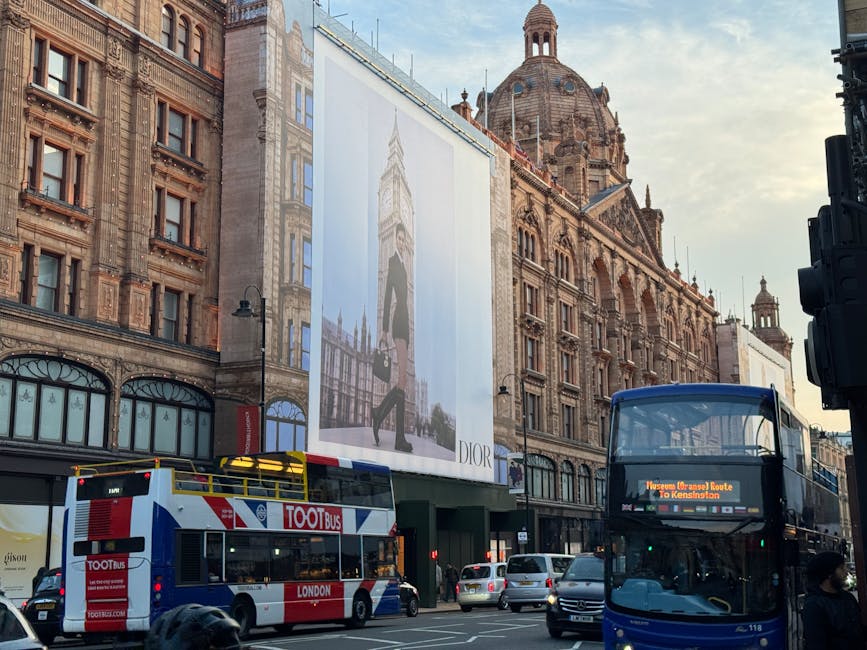 A city street scene in London showing historic architecture with ornate brick facades and a large domed building in the background. In the foreground, a double-decker bus with a digital route display 