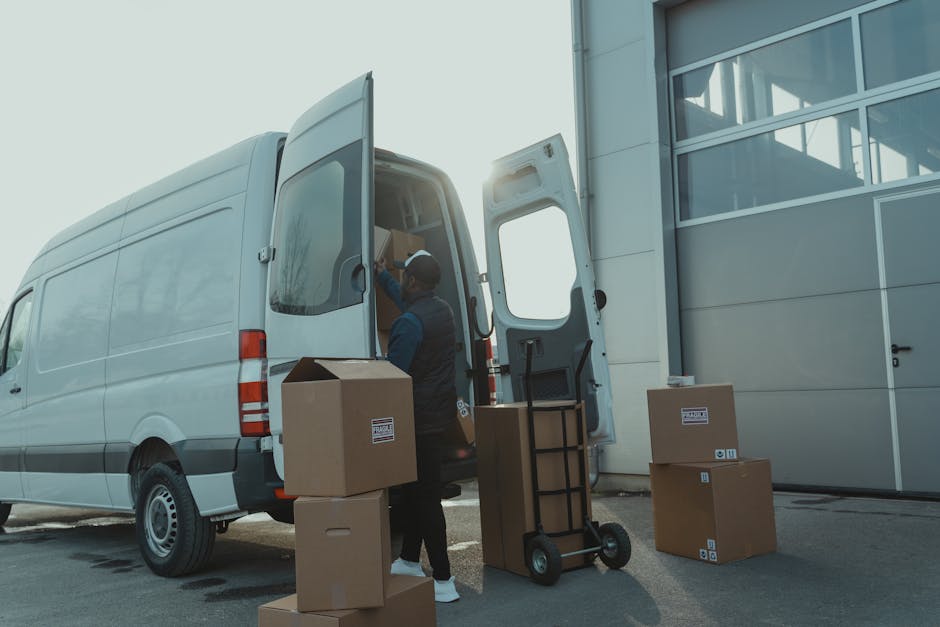 A professional mover from Knightsbridge Removals is loading packed cardboard boxes and various household items into a large white moving van parked outside a modern building with glass windows. The mo