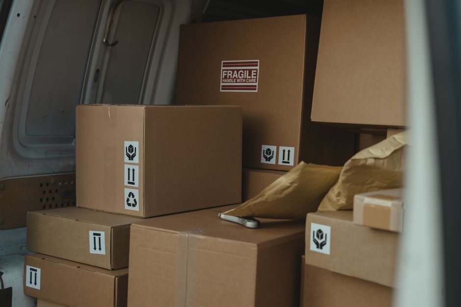 A professional mover from Knightsbridge Removals is loading packed cardboard boxes and various household items into a large white moving van parked outside a modern building with glass windows. The mo