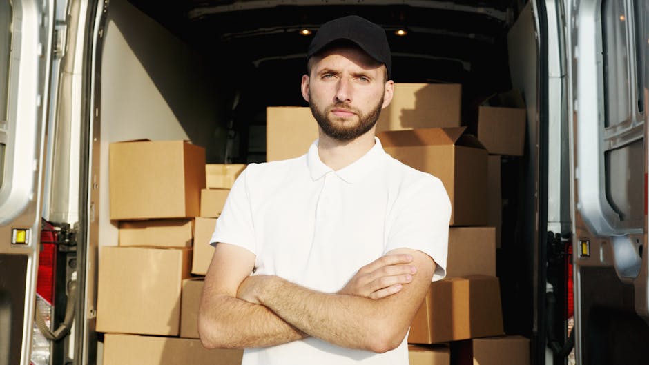 A young man with a serious expression, wearing a black cap and a white short-sleeved shirt, stands with arms crossed in front of an open moving van filled with packed cardboard boxes and wrapped furni