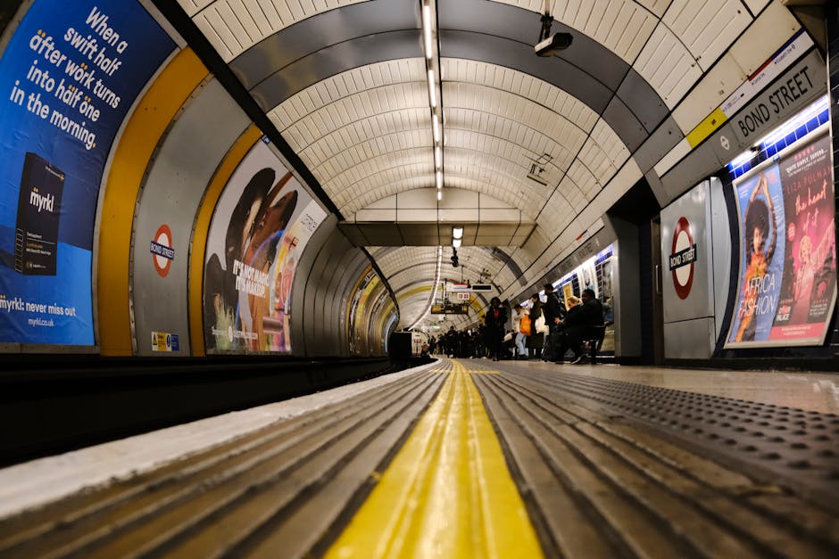 A wide-angle view of an underground London tube station platform, featuring a curved ceiling with white panels and overhead lighting. The platform has a yellow safety line running parallel to the edge