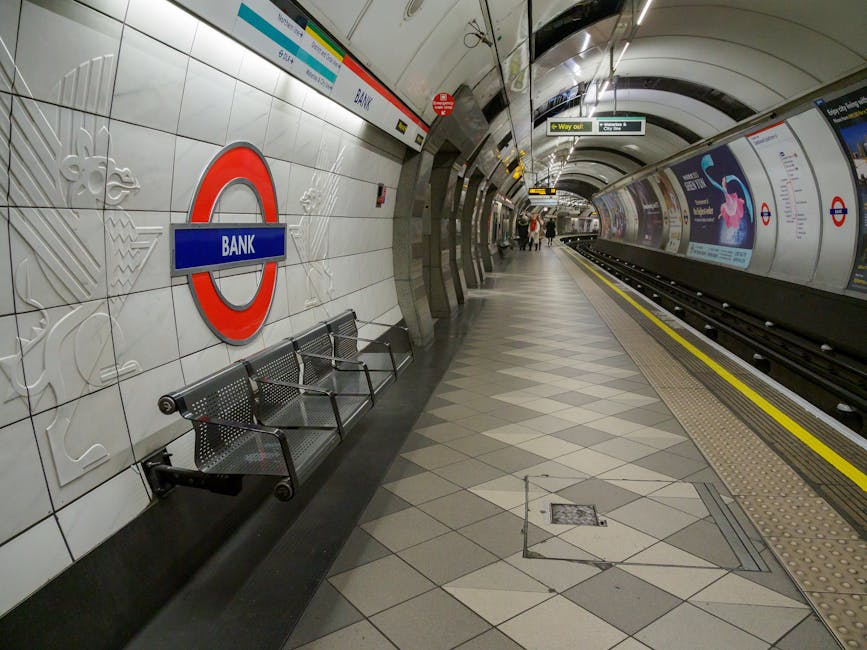 A wide-angle view of an underground London tube station platform, featuring a curved ceiling with white panels and overhead lighting. The platform has a yellow safety line running parallel to the edge