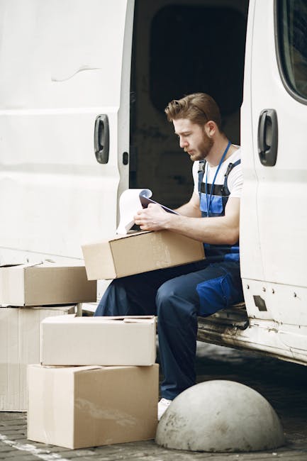 A man wearing a striped t-shirt and blue work overalls is seated at the open side door of a white van, surrounded by several cardboard boxes of various sizes, some stacked on the ground and others on 