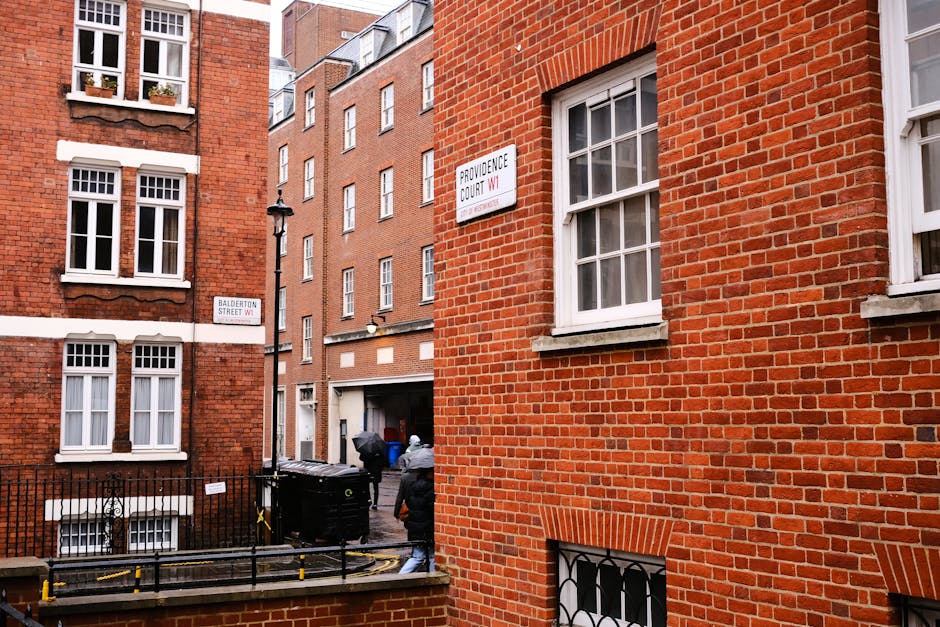 Photograph of a narrow residential street in Knightsbridge with red brick buildings and white-framed windows, showing an outdoor scene of a house removal process. Two individuals are seen outside a bu