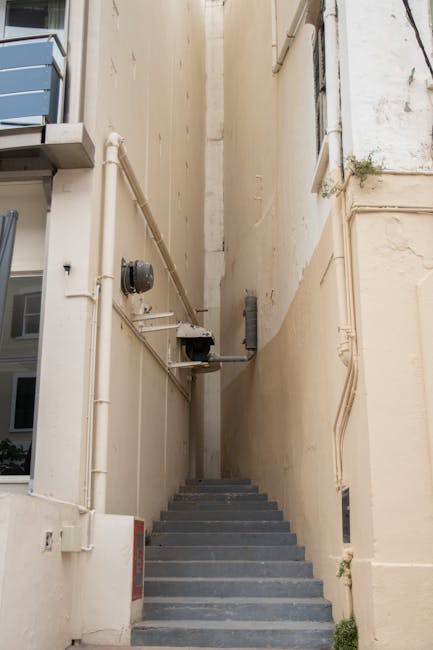 This image shows a narrow outdoor staircase located between two beige building walls, with multiple grey concrete steps leading upward. Along the left side of the staircase, there are several exposed 