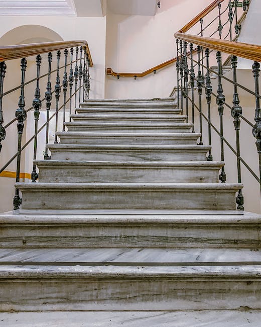 This image shows a narrow outdoor staircase located between two beige building walls, with multiple grey concrete steps leading upward. Along the left side of the staircase, there are several exposed 
